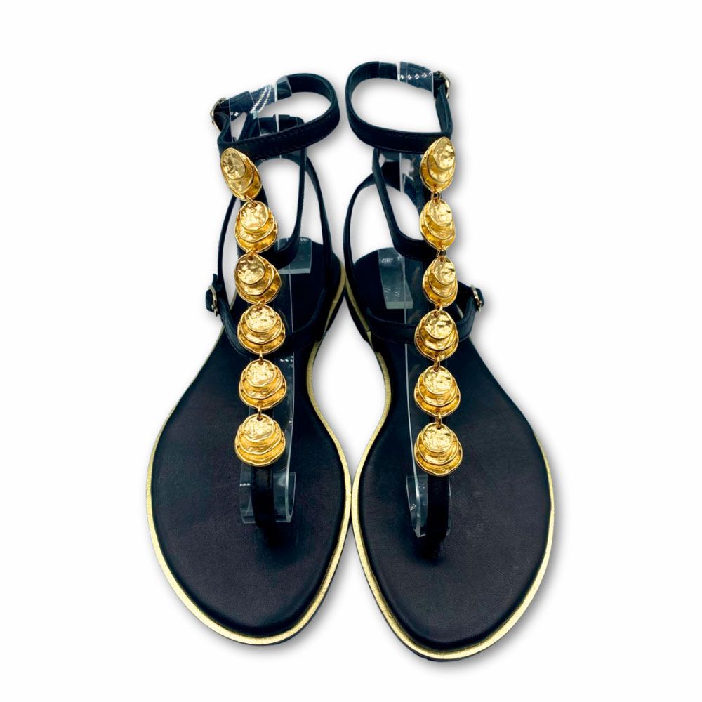 Sandalias griegas de piel color negro, con chapas doradas en tira central. Cierre con hebillas. Suela plana y tacón bajo de Exé.